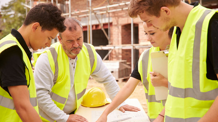 Three apprentices, including one woman, look at plans on a building site with a master builder.